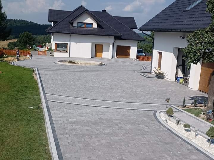Szeroki podjazd z szarej kostki brukowej wokół okrągłego klombu, z obrzeżami z ciemniejszej kostki, prowadzący do dwóch nowoczesnych domów z białymi elewacjami i ciemnymi dachami, w tle zalesione...