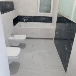 Nowoczesna łazienka w trakcie wykańczania: biała armatura (toaleta i bidet), wanna obudowana płytkami z geometrycznym wzorem, szare płytki podłogowe z subtelnym żyłkowaniem, fragment ściany...