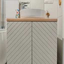 Nowoczesna łazienka z białą umywalką nablatową i szafką pod umywalką z szarymi drzwiczkami w jodełkę, chromowana bateria, minimalistyczny design, oświetlenie punktowe.