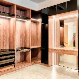 Nowoczesna szafa wnękowa z drewna z oświetleniem LED, czarnymi elementami i lustrem, widoczne wysuwane półki i kosze, logo producenta w odbiciu lustra.