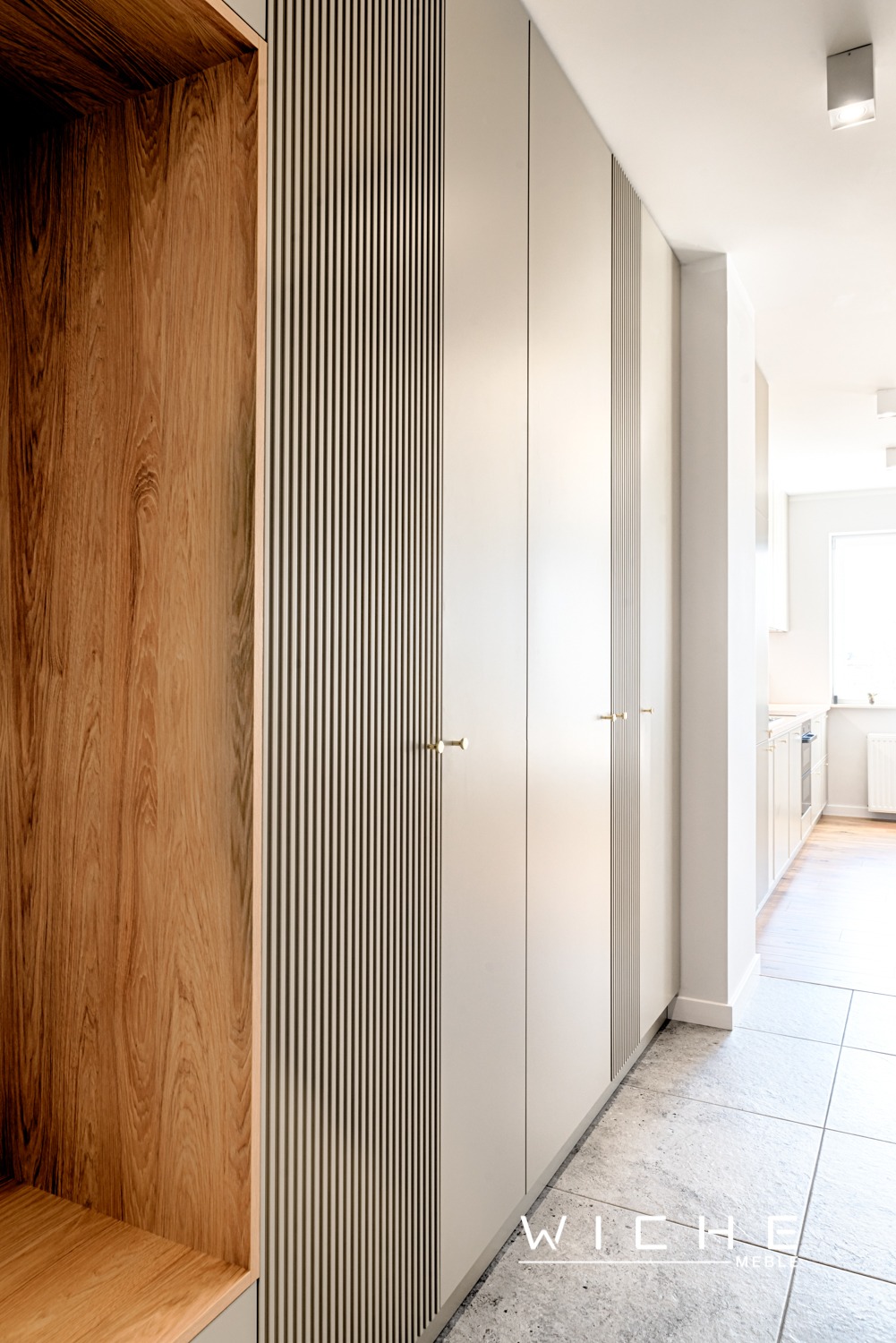 Wnętrze przedpokoju z zabudową meblową: szafa wnękowa z frontami w kolorze złamanej bieli, dekoracyjnymi pionowymi listwami i złotymi uchwytami, obok drewniany regał z widocznym usłojeniem.