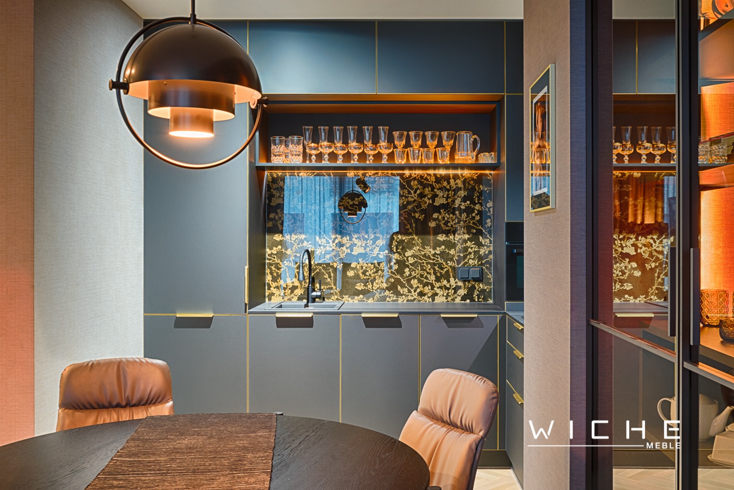 Elegancka, ciemnoszara kuchnia z złotymi akcentami, okrągły stół z dwukolorowym blatem i skórzane krzesła w aranżacji z ozdobnym oświetleniem i szkłem z motywem kwiatowym.