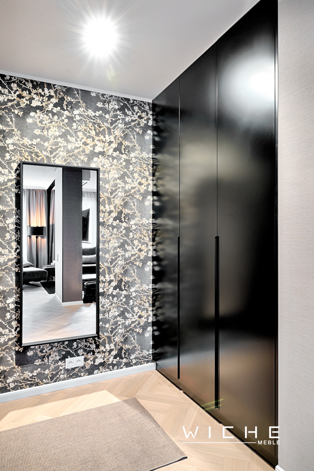 Wnętrze z dużą, czarną szafą wnękową z połyskiem, lustrem w czarnej ramie wiszącym na ścianie z tapetą w kwiaty, parkietem w jodełkę i beżowym dywanikiem.
