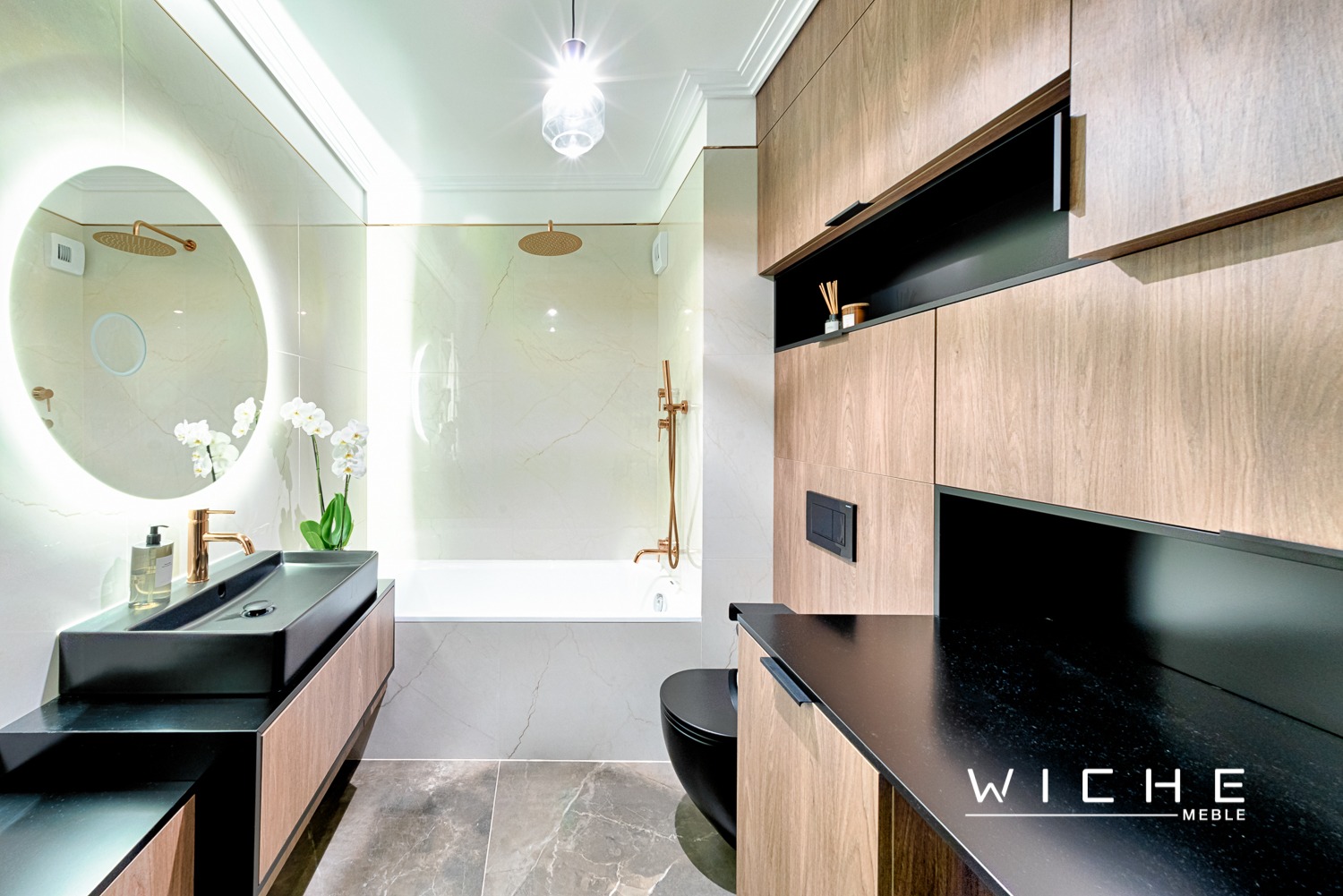 Nowoczesna łazienka z okrągłym lustrem podświetlanym LED, czarną umywalką nablatową, baterią w kolorze różowego złota i drewnianymi szafkami, uzupełniona białą wanną i czarną toaletą.