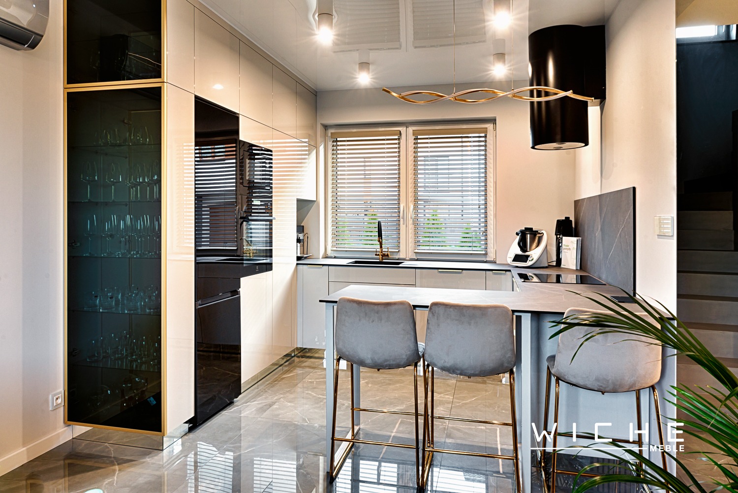 Nowoczesna, jasna kuchnia z wyspą, szarymi krzesłami barowymi, błyszczącymi frontami szafek i marmurową podłogą. Na blacie widoczny robot kuchenny i złota armatura.