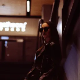 Kobieta w czarnej skórzanej kurtce i okularach przeciwsłonecznych, oparta o metalową ścianę z torebką z ćwiekami w dłoni, w tle neonowy napis.