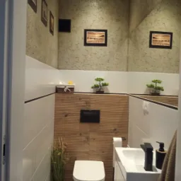 Nowoczesna, wykończona łazienka z beżowo-białymi płytkami, drewnianymi akcentami, lustrem na całej ścianie, toaletą, umywalką z czarną armaturą i dekoracyjnymi elementami, takimi jak sztuczne...