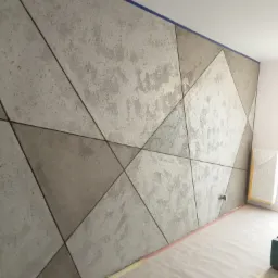 Ściana w pokoju pokryta dekoracyjną szarą strukturą z geometrycznym podziałem na trójkąty i romby, zabezpieczona taśmą malarską u góry i dołu, podłoga zakryta papierem malarskim.