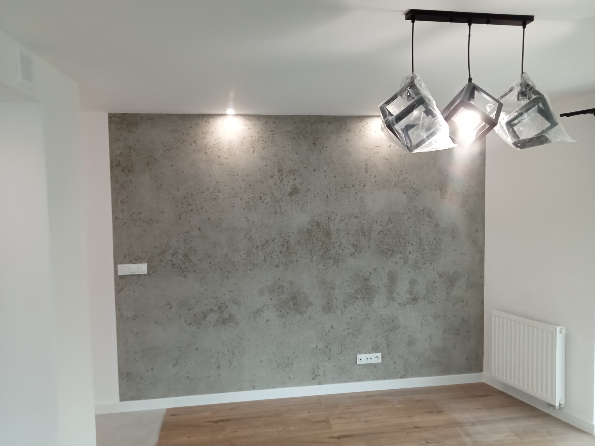 Ściana z efektem betonu architektonicznego w nowym mieszkaniu, z trzema lampami w geometrycznych abażurach, jeszcze w folii ochronnej.