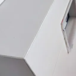 Biała, błyszcząca zabudowa z widocznym otworem technicznym i fragmentem ściany wyłożonej płytkami, sfotografowana pod kątem.