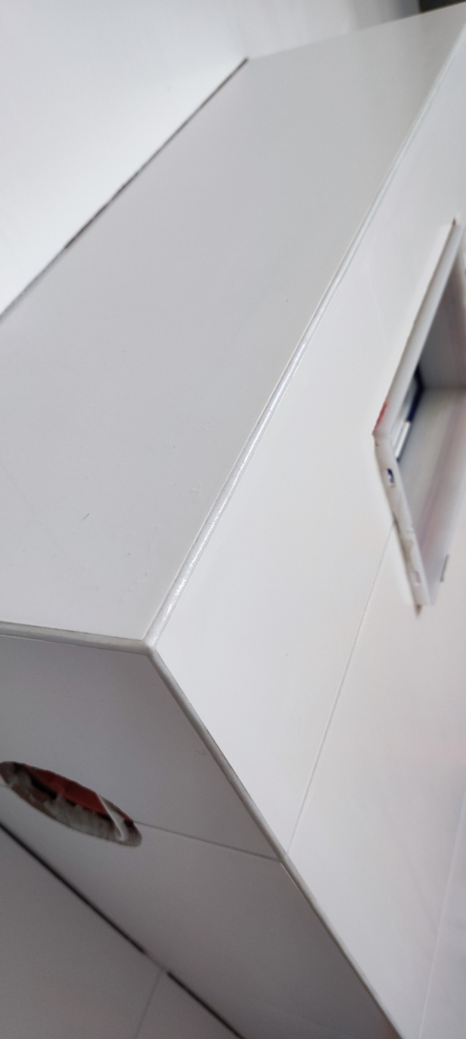 Biała, błyszcząca zabudowa z widocznym otworem technicznym i fragmentem ściany wyłożonej płytkami, sfotografowana pod kątem.