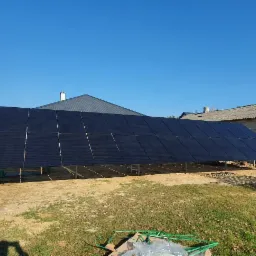 Panele słoneczne na konstrukcji wsporczej, widoczne zza białego samochodu dostawczego z numerem rejestracyjnym NO 2069T, w tle budynki mieszkalne i błękitne niebo.