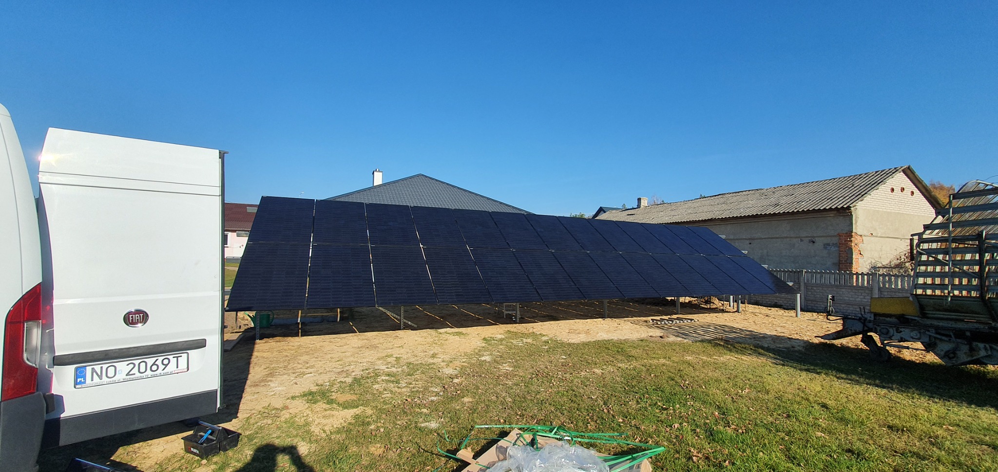 Panele słoneczne na konstrukcji wsporczej, widoczne zza białego samochodu dostawczego z numerem rejestracyjnym NO 2069T, w tle budynki mieszkalne i błękitne niebo.