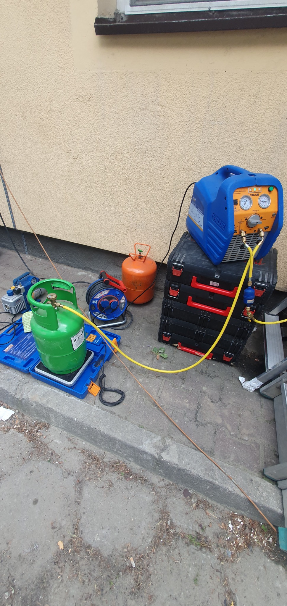 Sprzęt do odzyskiwania czynnika chłodniczego, butle z gazem R134a i narzędzia na chodniku przy ścianie budynku.