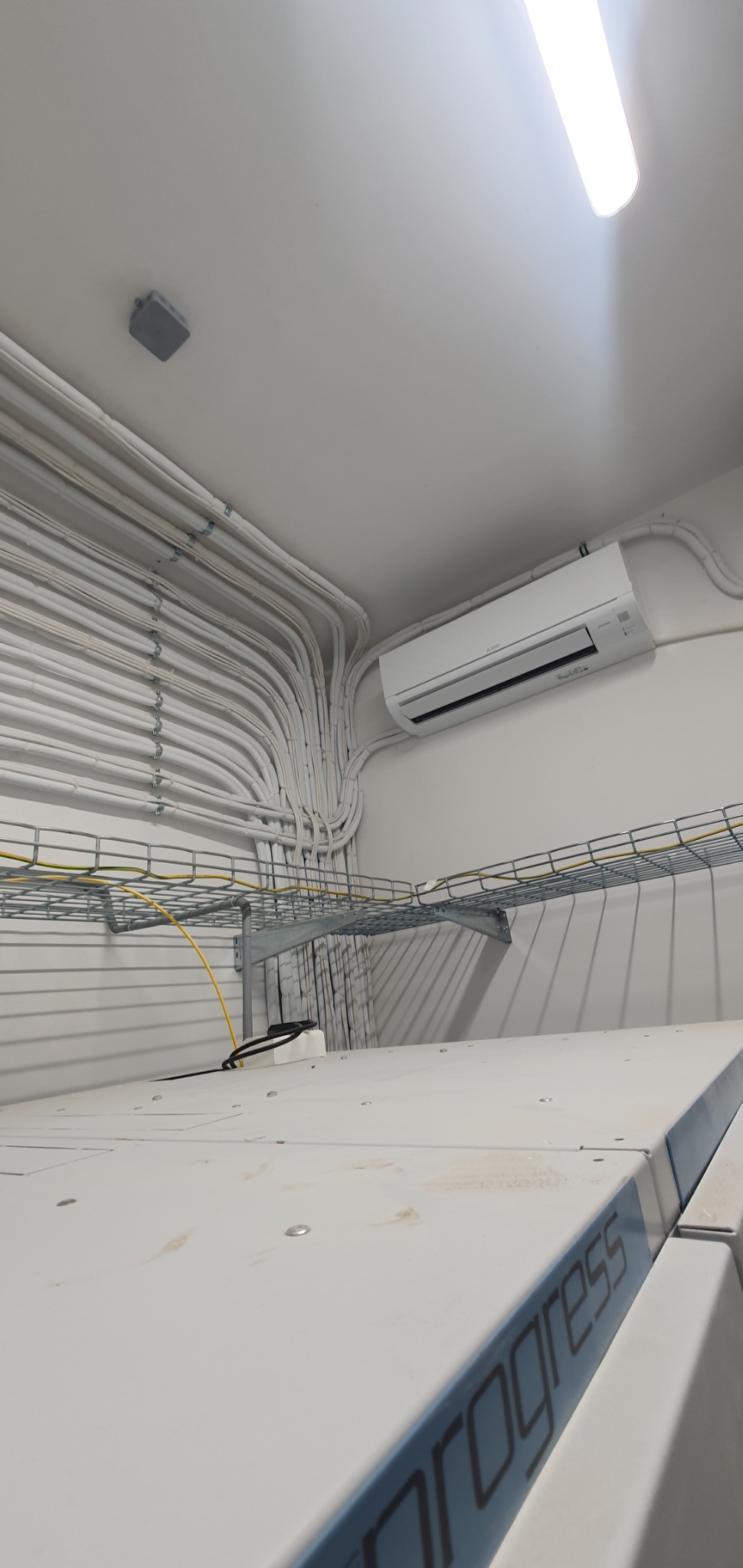 Wewnętrzna jednostka klimatyzacji zamontowana na ścianie, widoczne liczne peszle elektryczne prowadzone po suficie i ścianie, metalowa drabinka kablowa pod sufitem, fragment urządzenia z napisem...