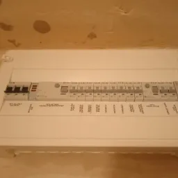 Rozdzielnica elektryczna natynkowa z widocznymi bezpiecznikami i opisami obwodów: gniazda pokoju, łazienki, lodówki, pralki, indukcji, piekarnika, zmywarki, oświetlenie, zasilanie dzwonka.