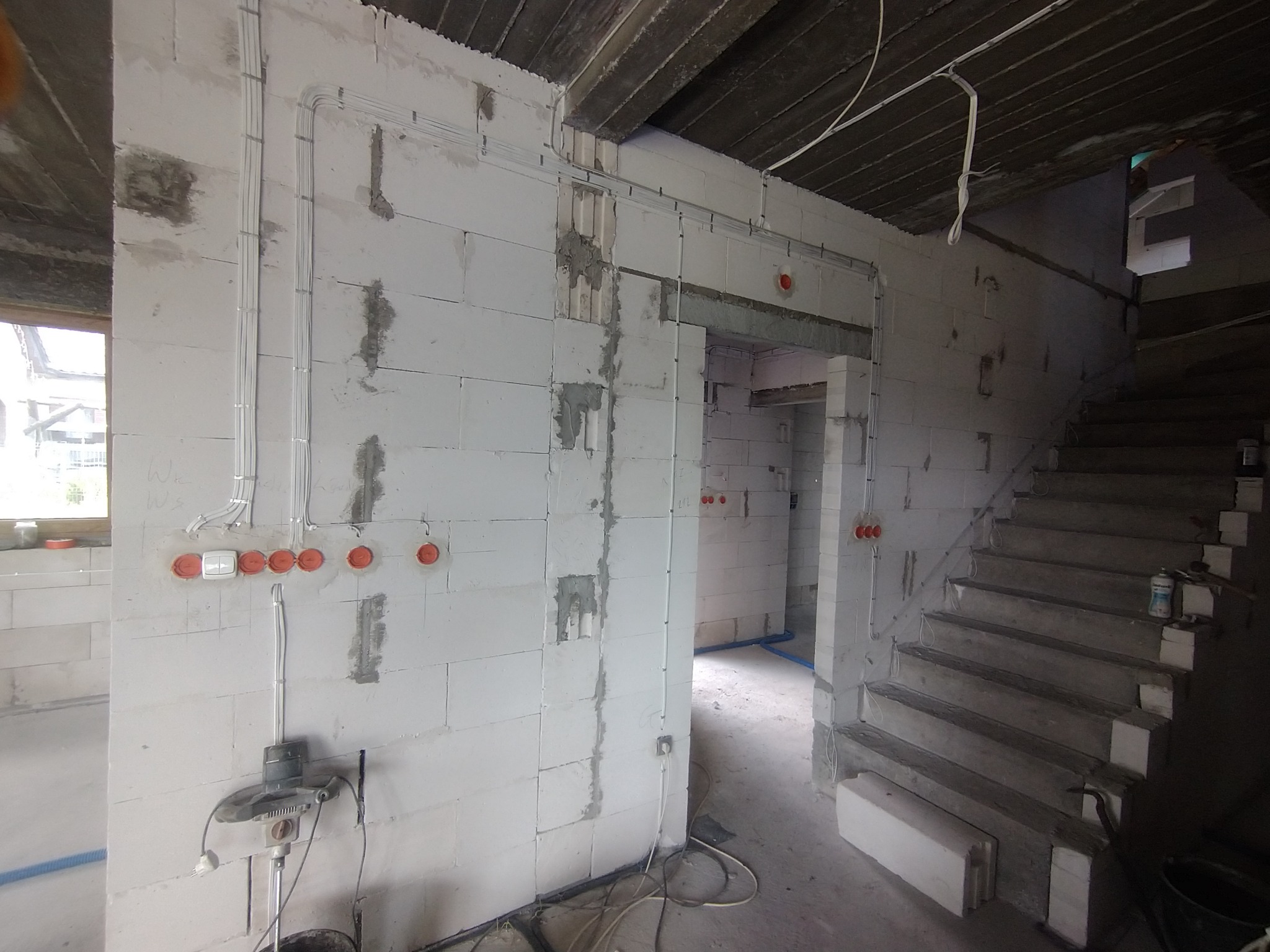 Wewnętrzne ściany budynku w trakcie budowy z widocznymi przewodami elektrycznymi w peszlach, puszkami instalacyjnymi, oraz narzędziem do wiercenia otworów pod gniazdka, surowe schody betonowe w tle.