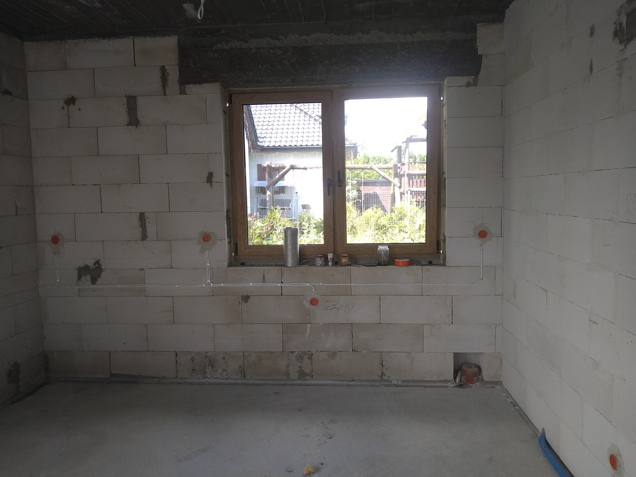 Surowy stan wnętrza w trakcie prac: ściany z białych bloczków, okno z widokiem na ogród, widoczne przewody elektryczne w peszlach z puszkami instalacyjnymi, fragment instalacji podtynkowej w bruździe.