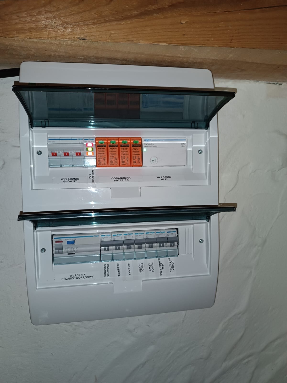 Rozdzielnica elektryczna Hager z otwartymi klapkami, widoczne wyłączniki, ograniczniki przepięć i włącznik WiFi. Na górnym panelu świecą się diody, wskazując status obwodów. Ściana o nierównej...