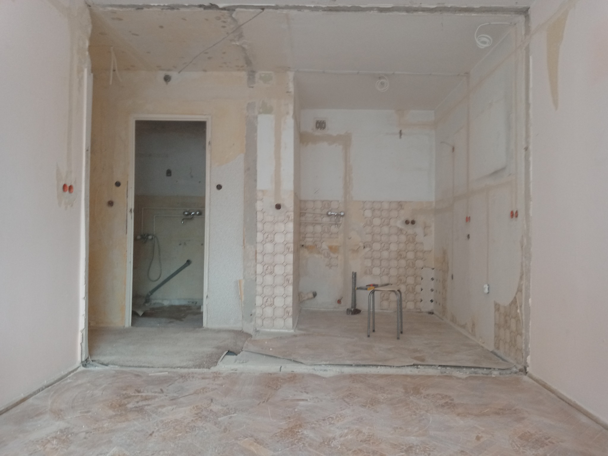 Remont mieszkania w trakcie: zerwane tapety, odsłonięte instalacje elektryczne, widoczne puszki elektryczne, krzesło i narzędzia w pomieszczeniu, widok na łazienkę w tle.