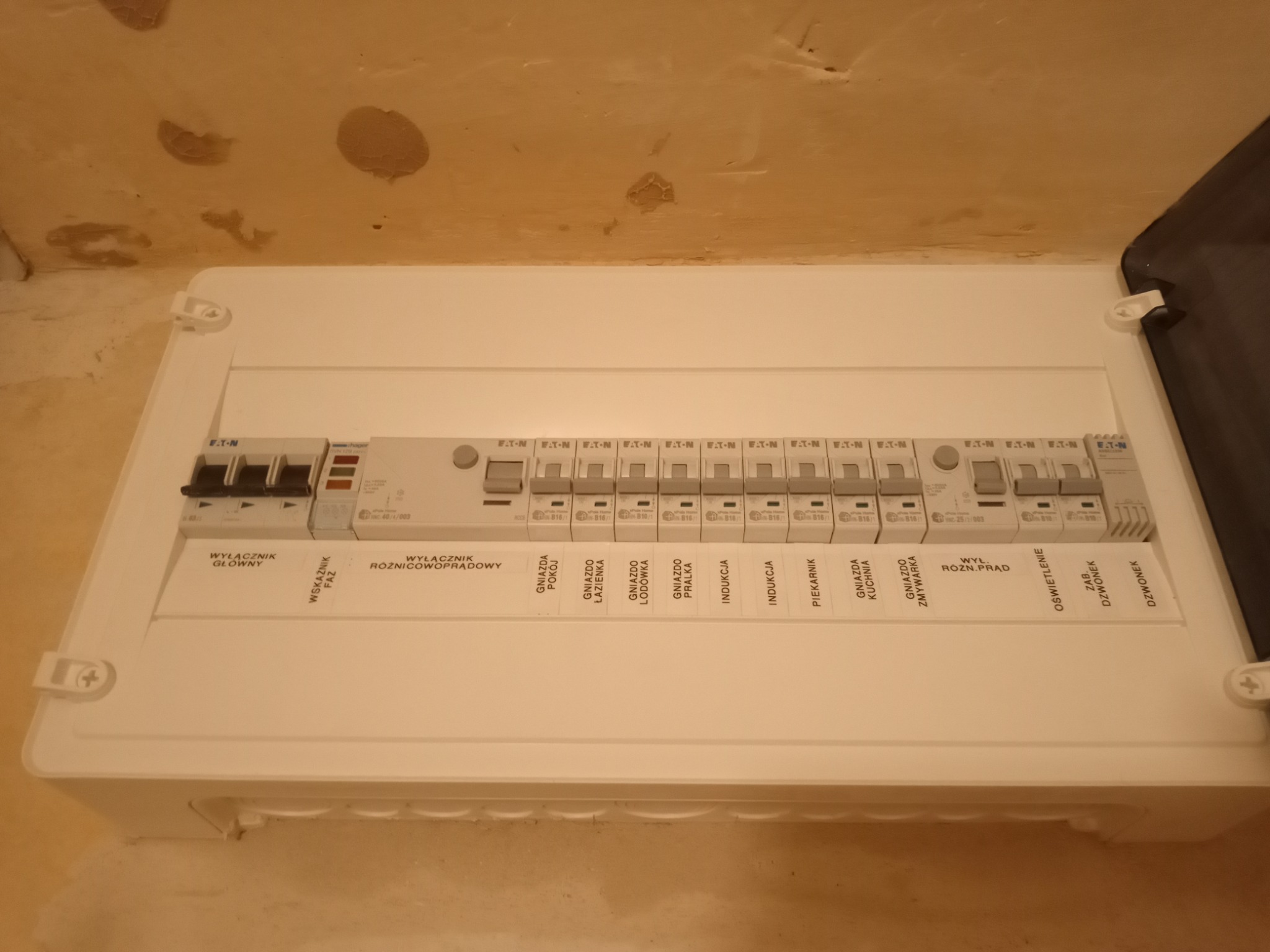 Rozdzielnica elektryczna natynkowa z widocznymi bezpiecznikami i opisami obwodów: gniazda pokoju, łazienki, lodówki, pralki, indukcji, piekarnika, zmywarki, oświetlenie, zasilanie dzwonka.