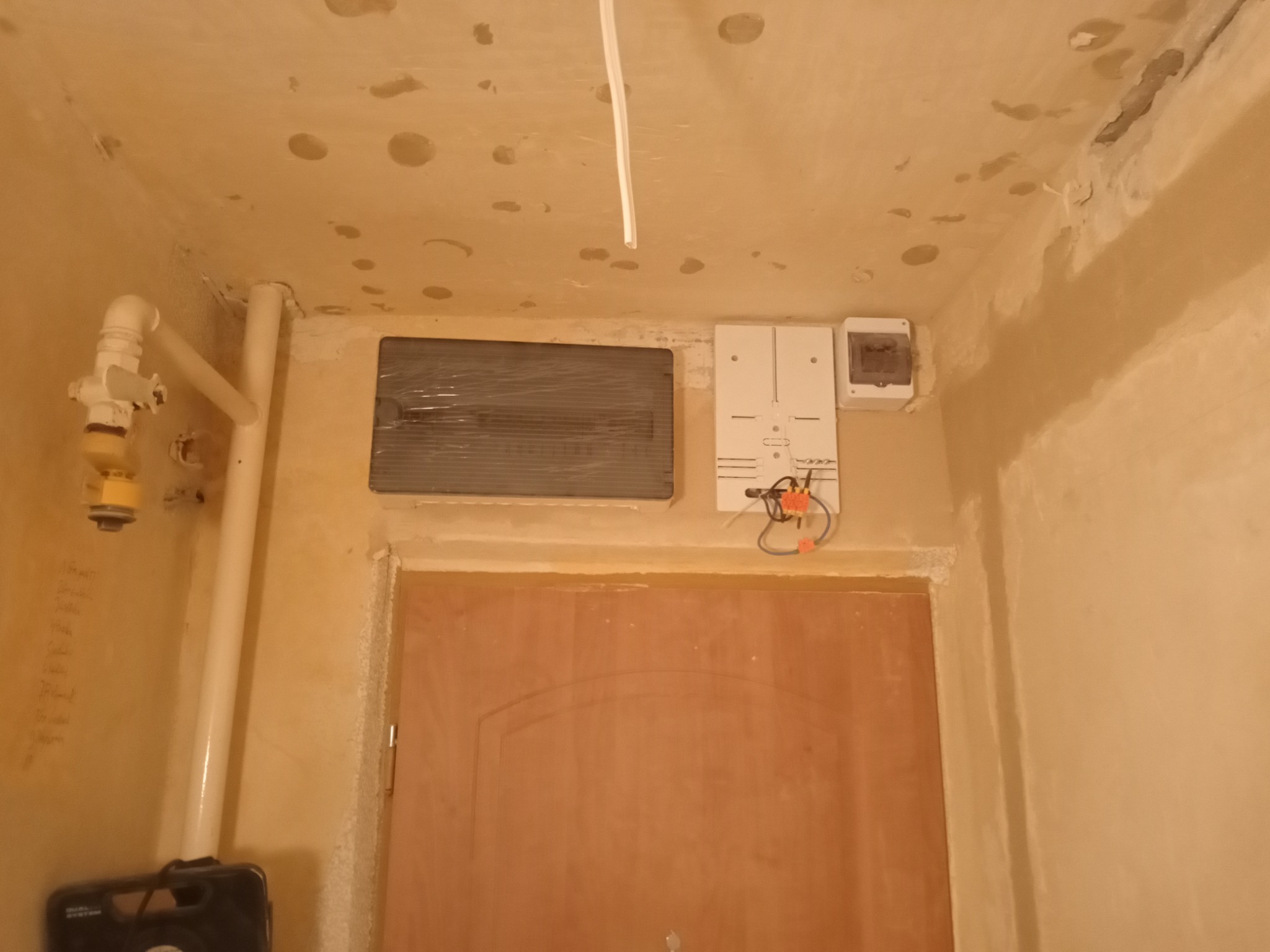 Niezakończone prace elektryczne: nowa skrzynka bezpiecznikowa i podłączenia przewodów nad futryną drzwi w surowym wnętrzu, widoczne rury instalacyjne i niedoskonałości na ścianach i suficie.