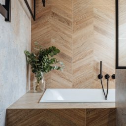 Project-art - Nowoczesna łazienka z wanną obudowaną płytkami imitującymi drewno ułożonymi w jodełkę, czarna armatura i wazon z eukaliptusem.