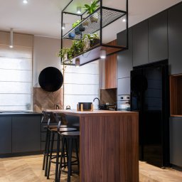 Project-art - Nowoczesna kuchnia z wyspą z ciemnego drewna, czarnymi szafkami i podłogą w jodełkę, oświetlona lampami punktowymi i z metalową konstrukcją z roślinami nad wyspą.