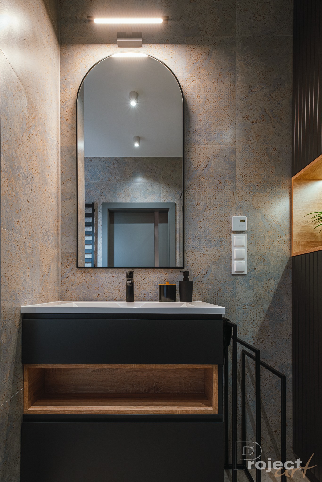 Nowoczesna łazienka z umywalką nablatową, czarną armaturą i lustrem w kształcie łuku, podświetlonym od góry, na tle płytek w odcieniach szarości i beżu z delikatnym wzorem.