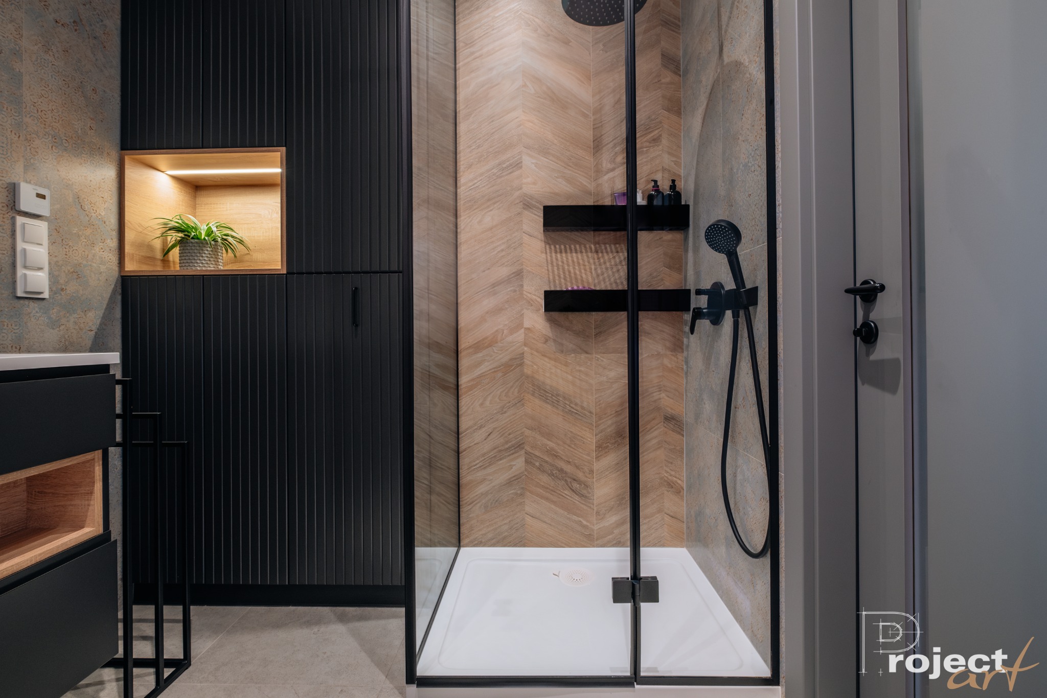 Nowoczesna łazienka z kabiną prysznicową typu walk-in, wykończona płytkami w jodełkę, czarnymi meblami z ryflowanymi frontami i oświetloną wnęką z rośliną.