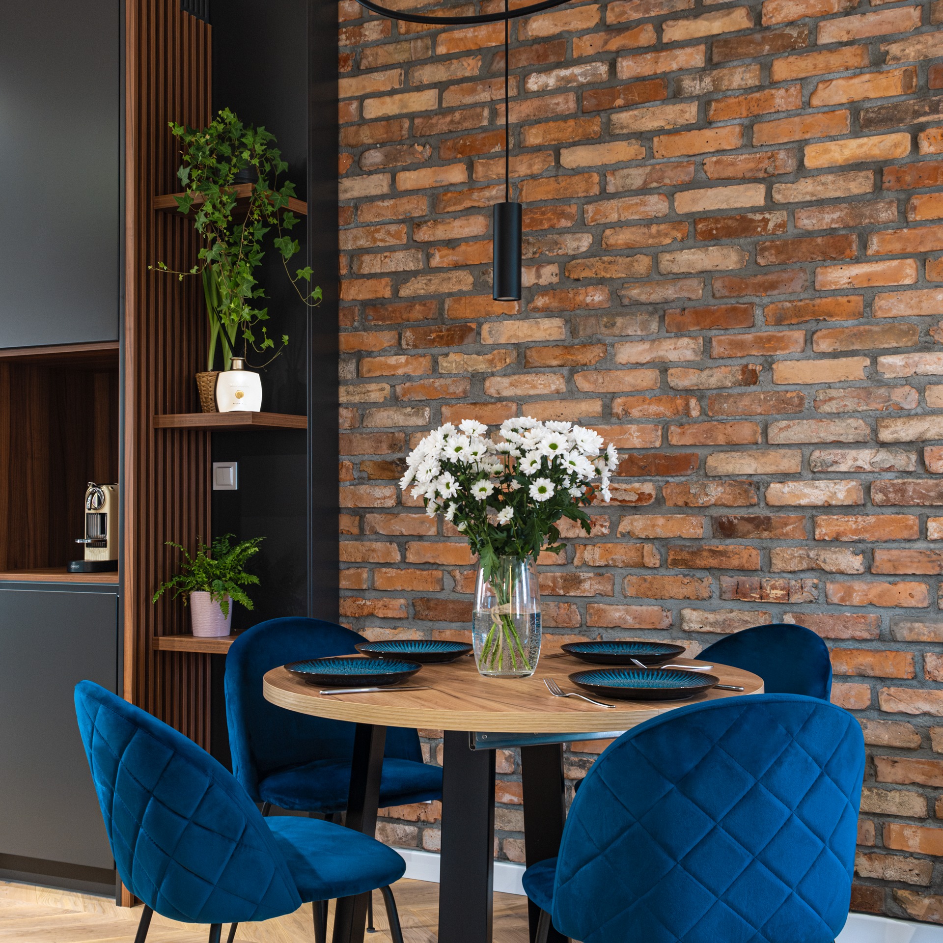 Jadalnia z okrągłym drewnianym stołem, nakrytym na cztery osoby, otoczonym przez cztery granatowe, welurowe krzesła na tle ściany z czerwonej cegły i zabudowy kuchennej z drewnianymi lamelami...