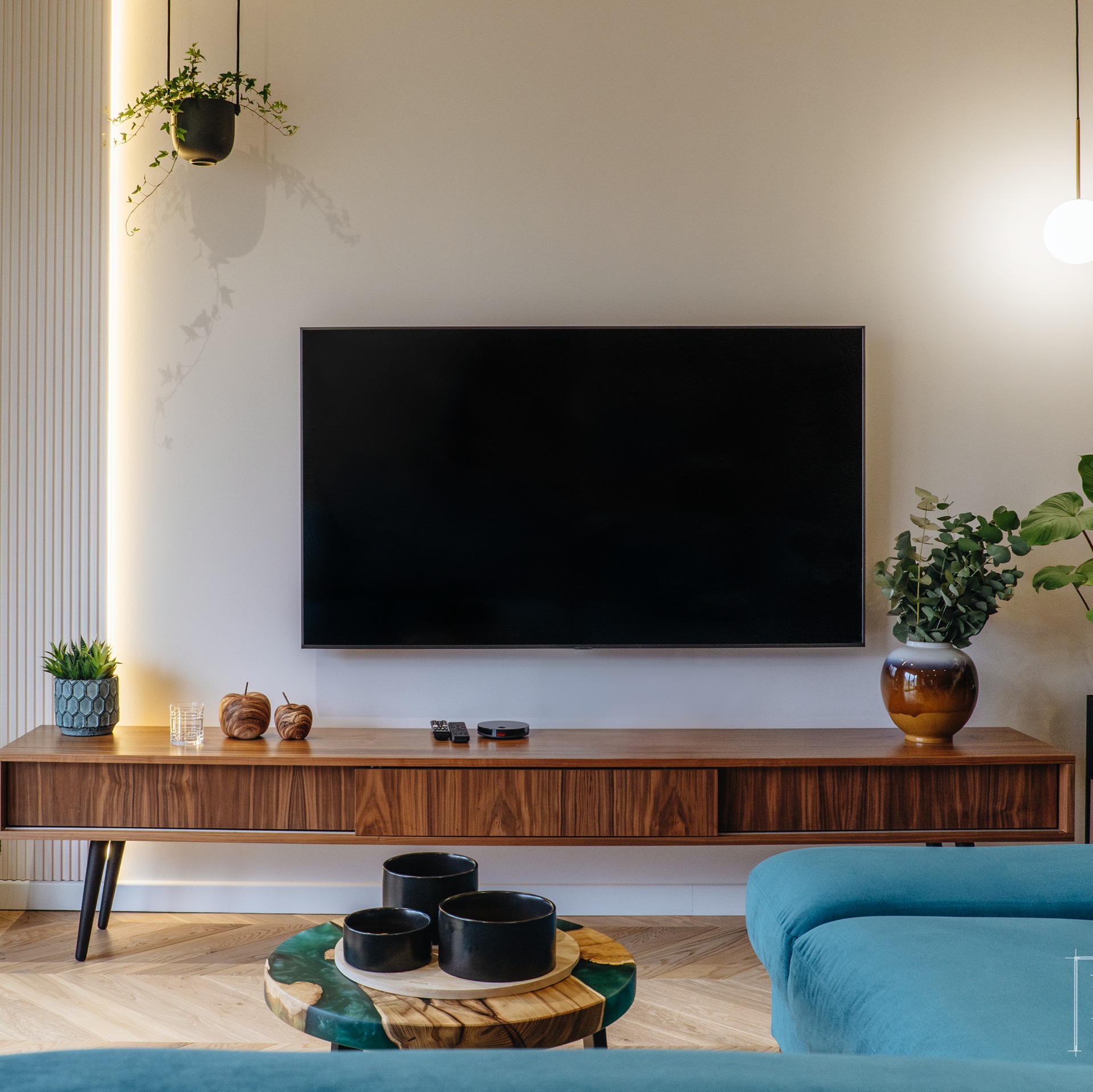 Salon z telewizorem na drewnianej szafce RTV, okrągły stolik kawowy z żywicą epoksydową, turkusowa sofa modułowa, parkiet w jodełkę i dekoracje roślinne.