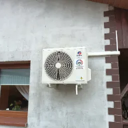 Zewnętrzna jednostka klimatyzacji marki Gree zamontowana na szarej, betonowej ścianie budynku z widocznym oknem i dekoracyjnym wykończeniem z cegły.