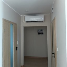 KLIMAT Mateusz Wiluk - Wnętrze mieszkania z zamontowanym klimatyzatorem typu split marki AUX, ustawionym na 23 stopnie, widocznym nad wejściem do kolejnego pomieszczenia z obrazem na ścianie, z perspektywy korytarza...