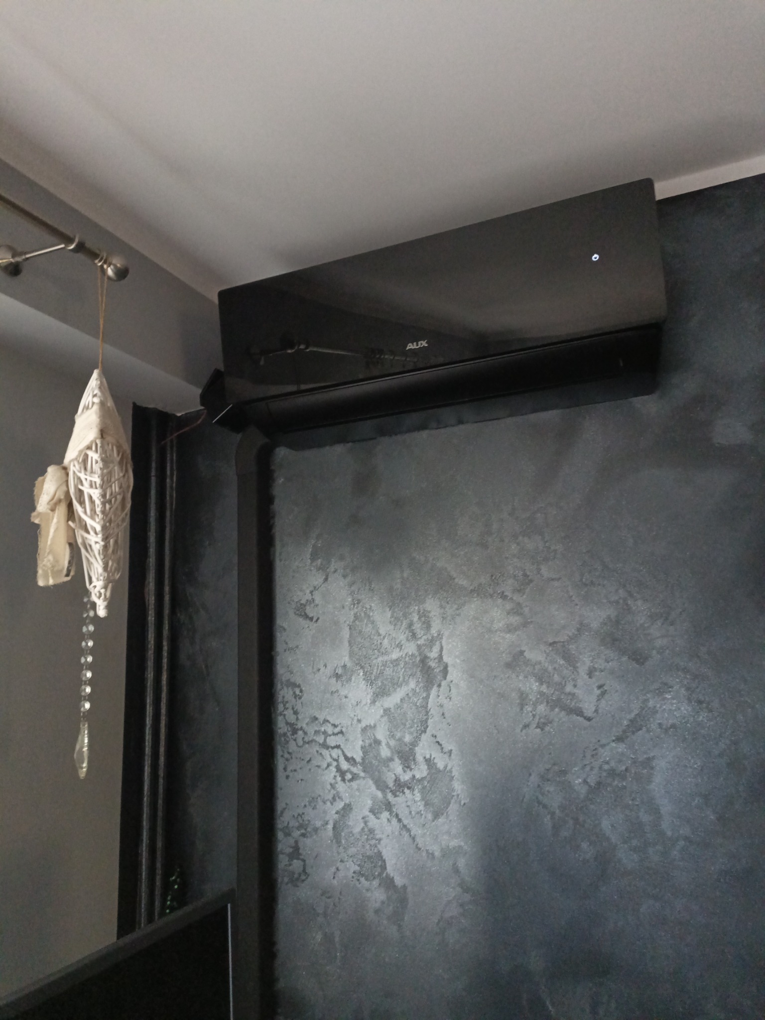 Czarny klimatyzator marki AUX zamontowany na ścianie o dekoracyjnej, szarej fakturze, z widoczną ozdobą wiszącą w lewym górnym rogu.