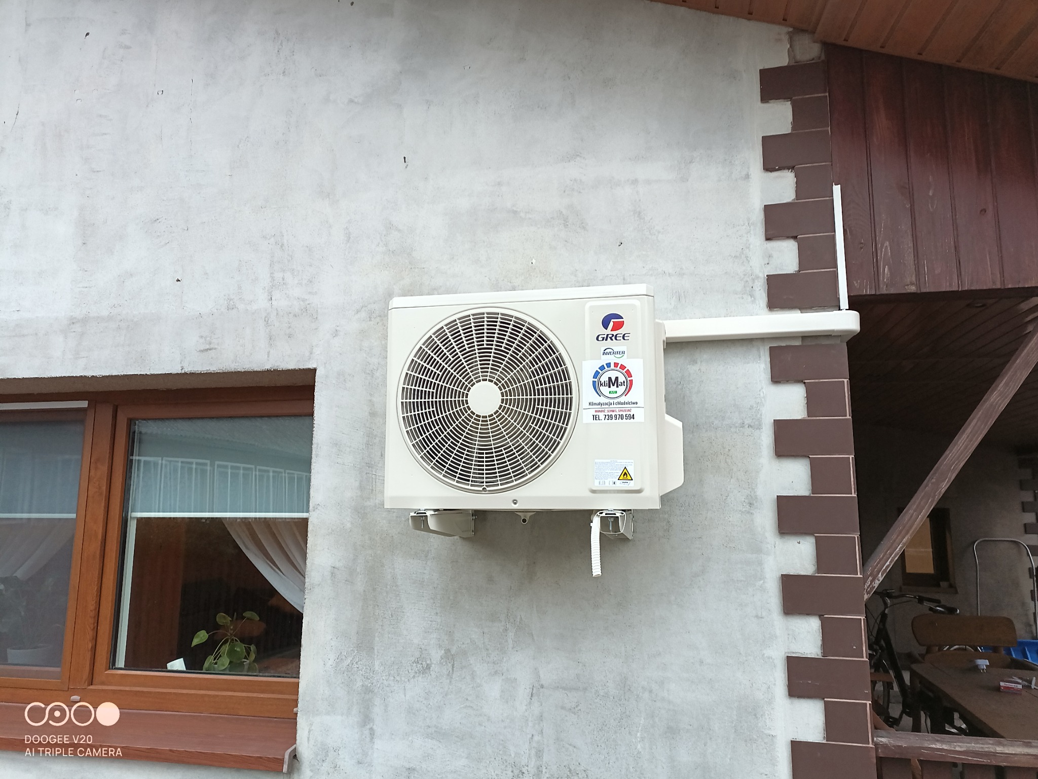 Zewnętrzna jednostka klimatyzacji marki Gree zamontowana na szarej, betonowej ścianie budynku z widocznym oknem i dekoracyjnym wykończeniem z cegły.