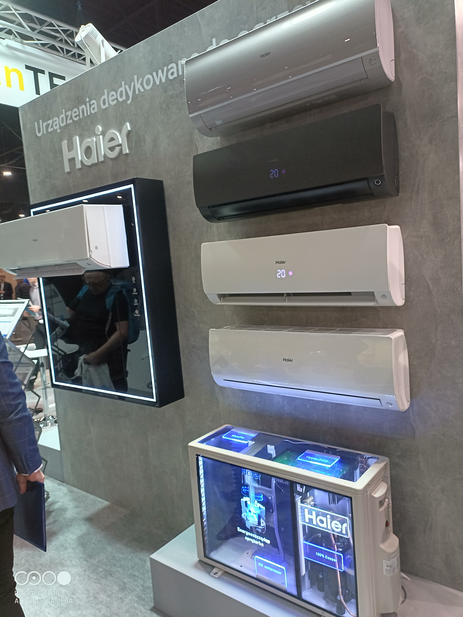 Ekspozycja klimatyzatorów Haier w różnych kolorach (biały, szary, czarny) na ścianie, z modelem demonstracyjnym z przezroczystą obudową prezentującym wewnętrzne komponenty, widoczne hasło...