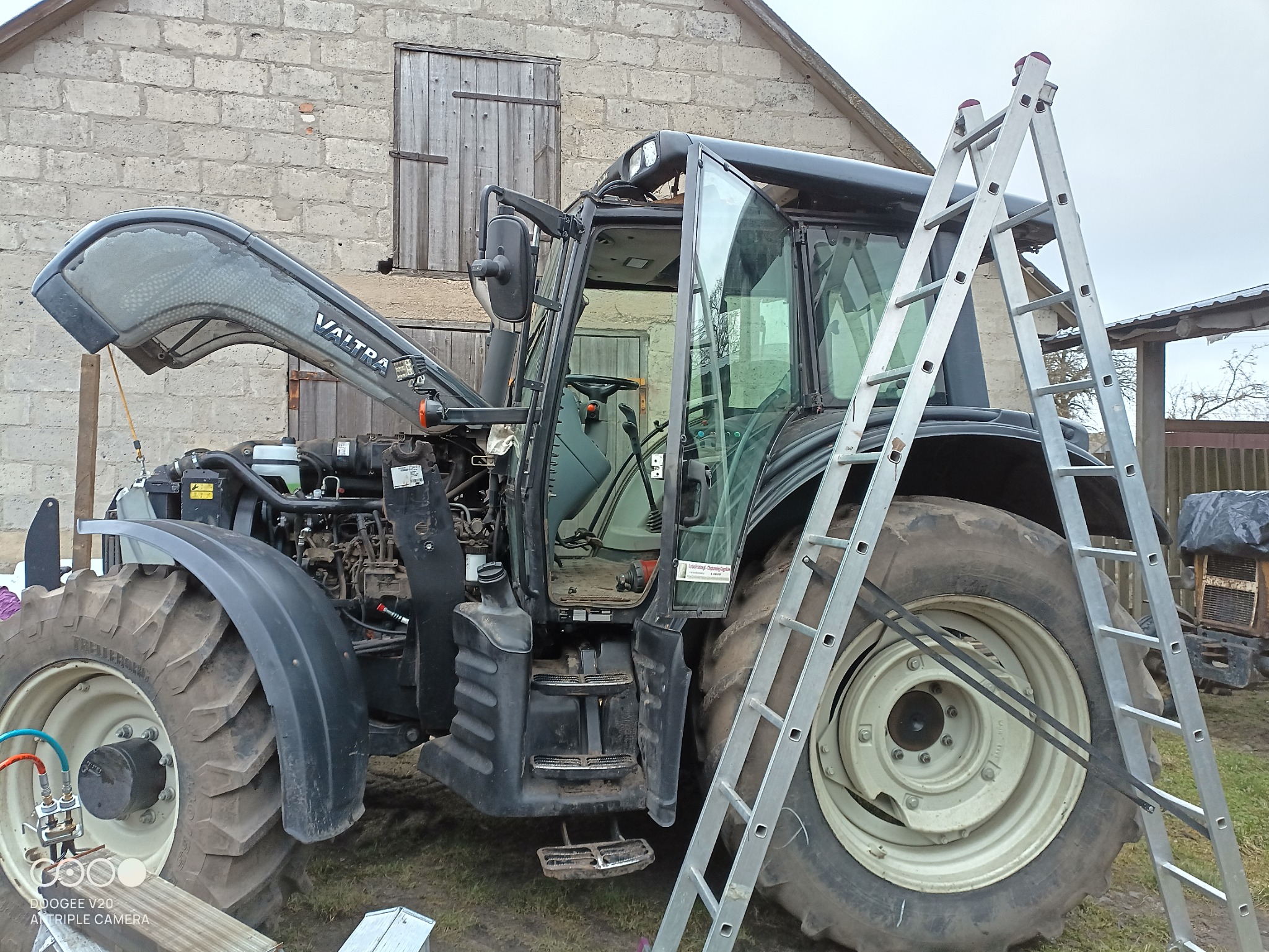 Otwarta maska silnika ciągnika Valtra, widoczny silnik, obok drabina aluminiowa oparta o koło, tło stanowi murowany budynek gospodarczy z drewnianymi drzwiami.