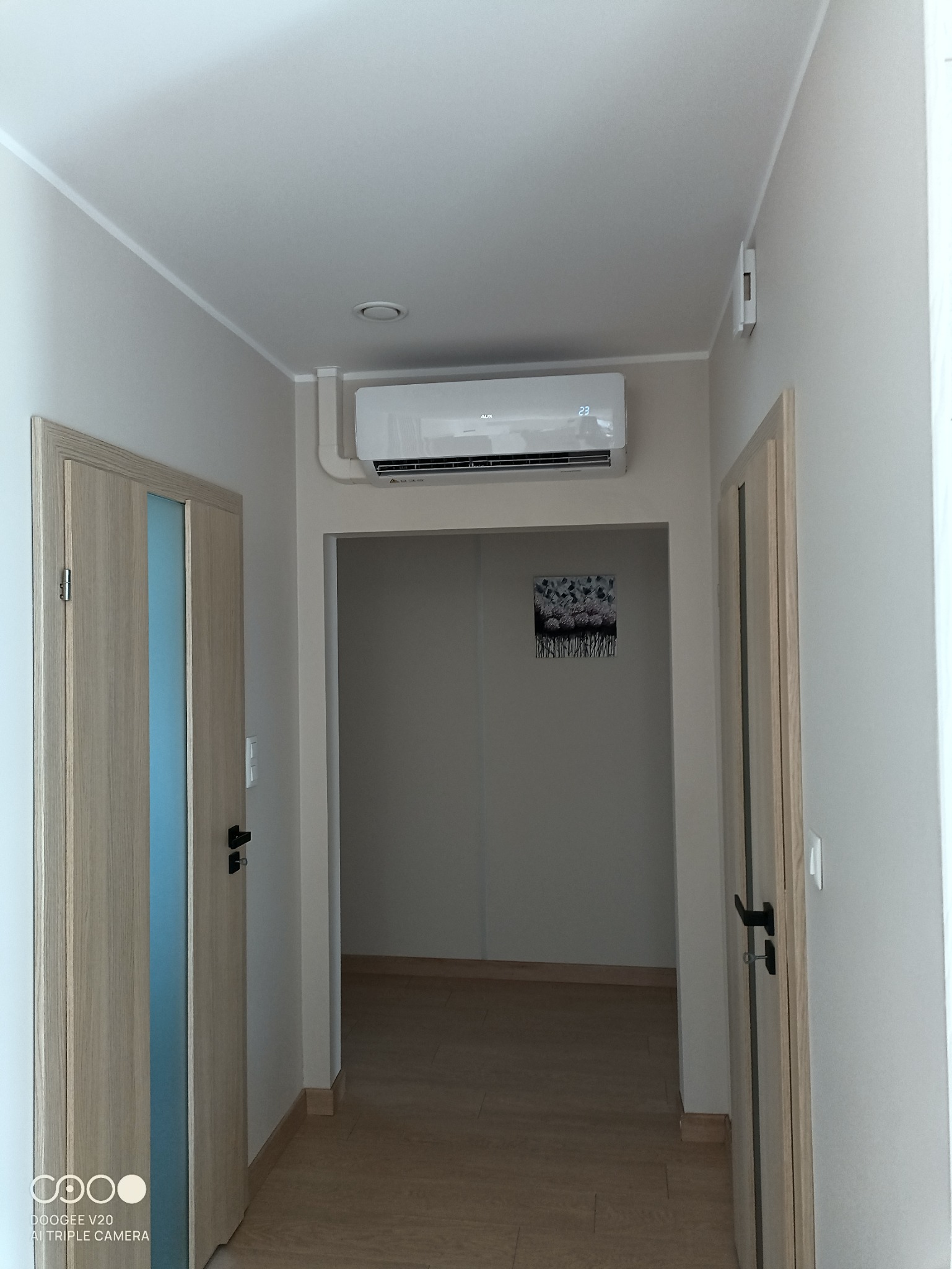 Wnętrze mieszkania z zamontowanym klimatyzatorem typu split marki AUX, ustawionym na 23 stopnie, widocznym nad wejściem do kolejnego pomieszczenia z obrazem na ścianie, z perspektywy korytarza...