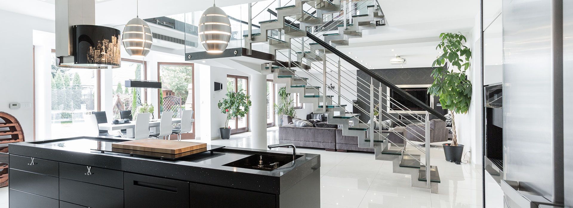 Wnętrze przestronnego, nowoczesnego domu z otwartą kuchnią z czarną wyspą i drewnianą deską do krojenia, schodami z metalu i szkła oraz salonem z szarą sofą w tle.