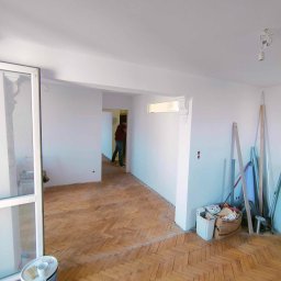 Przedsiębiorstwo Usługowe Jarosław Butowski - Pokój po malowaniu ścian na biało, z parkietem w jodełkę, widoczne ślady po remoncie, puszka z farbą i kubek na podłodze, w tle osoba przechodząca do kolejnego pomieszczenia.