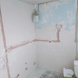 Przedsiębiorstwo Usługowe Jarosław Butowski - Wnętrze w trakcie prac remontowych: ściany częściowo pokryte gładzią, widoczne instalacje elektryczne i rury, na podłodze worek z zaprawą.