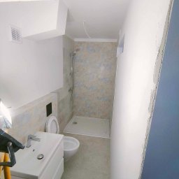 Przedsiębiorstwo Usługowe Jarosław Butowski - Wnętrze łazienki po remoncie: biała umywalka z szafką, toaleta podwieszana, kabina prysznicowa z jasnymi płytkami z wzorem, wentylator w suficie, fragment ściany z niedokończonymi gładziami.