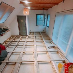 Przedsiębiorstwo Usługowe Jarosław Butowski - Poddasze w trakcie remontu podłogi: drewniana konstrukcja legarów ułożona na surowym podłożu, widoczna osoba w czerwonej koszuli w kratę, okno dachowe zasłonięte folią, lampa z wikliny.