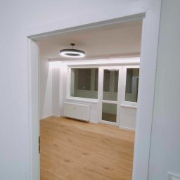 Przedsiębiorstwo Usługowe Jarosław Butowski - Pusty, jasny pokój po remoncie z drewnianą podłogą, białymi ścianami i nowoczesną okrągłą lampą sufitową, widok zza futryny drzwi.