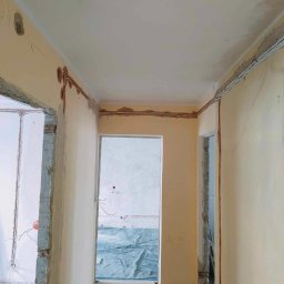 Przedsiębiorstwo Usługowe Jarosław Butowski - Korytarz w trakcie prac remontowych: odkryte przewody elektryczne w bruzdach ściennych, przygotowane do zalania zaprawą, surowy stan ścian przed wykończeniem.