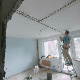 Przedsiębiorstwo Usługowe Jarosław Butowski - Mężczyzna na drabinie szpachluje sufit w pomieszczeniu w trakcie remontu, widoczne kable elektryczne, worki z zaprawą i uszkodzone fragmenty ścian.