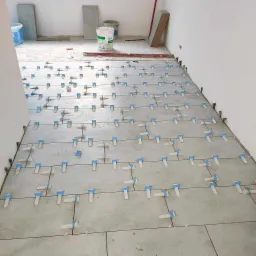 Układanie jasnoszarych płytek podłogowych z użyciem systemu poziomowania płytek z niebieskimi klinami, widoczne wiadro z zaprawą i poziomica na podłodze w tle.