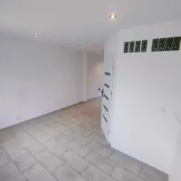 Pusty, odnowiony pokój z szarymi płytkami podłogowymi ułożonymi w prostokąty, białe ściany, oświetlenie punktowe w suficie i częściowo otwarte białe drzwi z ciemnymi paskami.