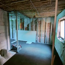 Przedsiębiorstwo Usługowe Jarosław Butowski - Pomieszczenie w trakcie remontu: surowa cegła, metalowa konstrukcja, płyta kartonowo-gipsowa oparta o ścianę, narzędzia w skrzynce.